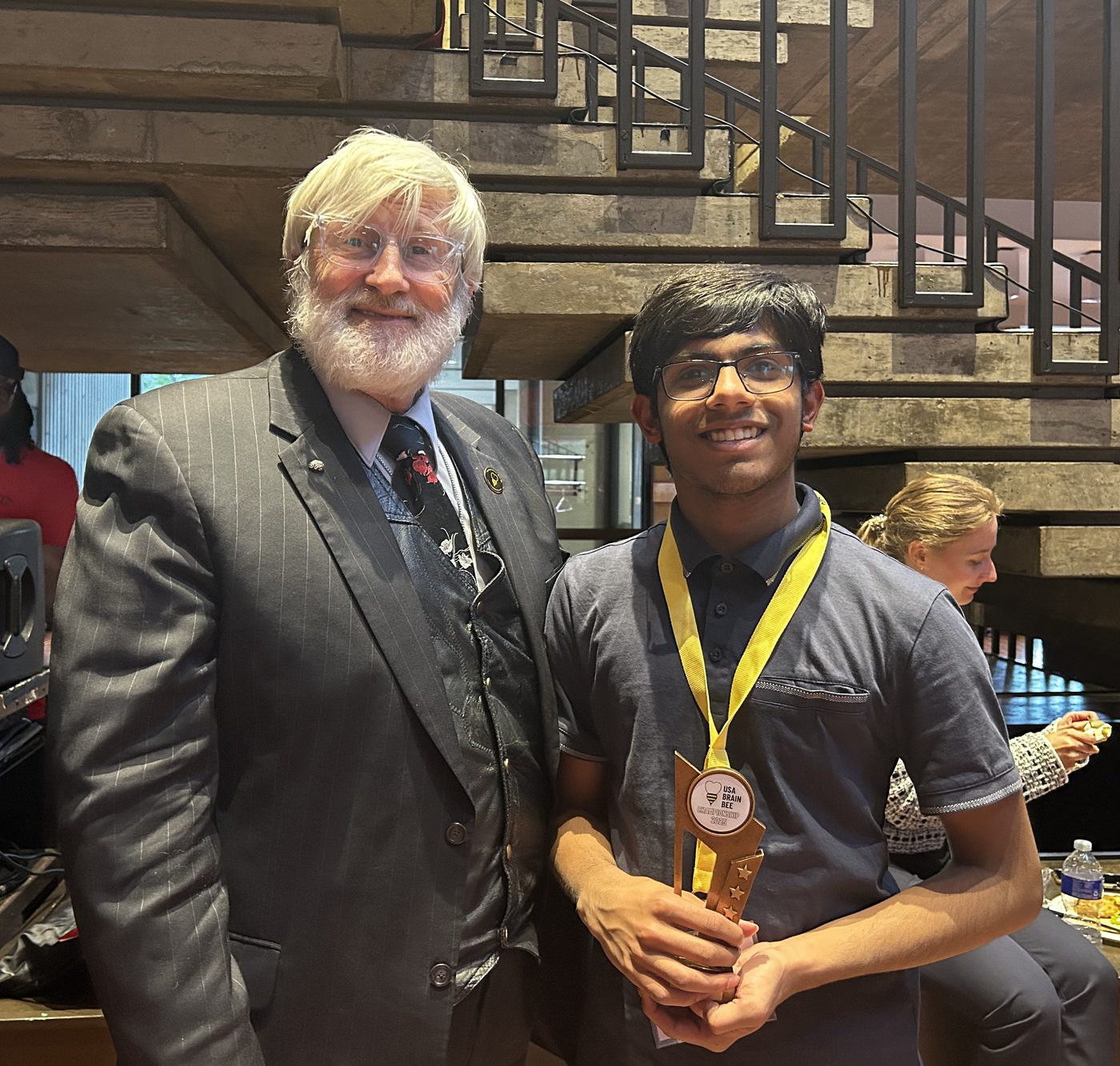 Navneeth Badhri holding the USA Brain Bee trophy at the national finals, standing next to the competition's organizer.